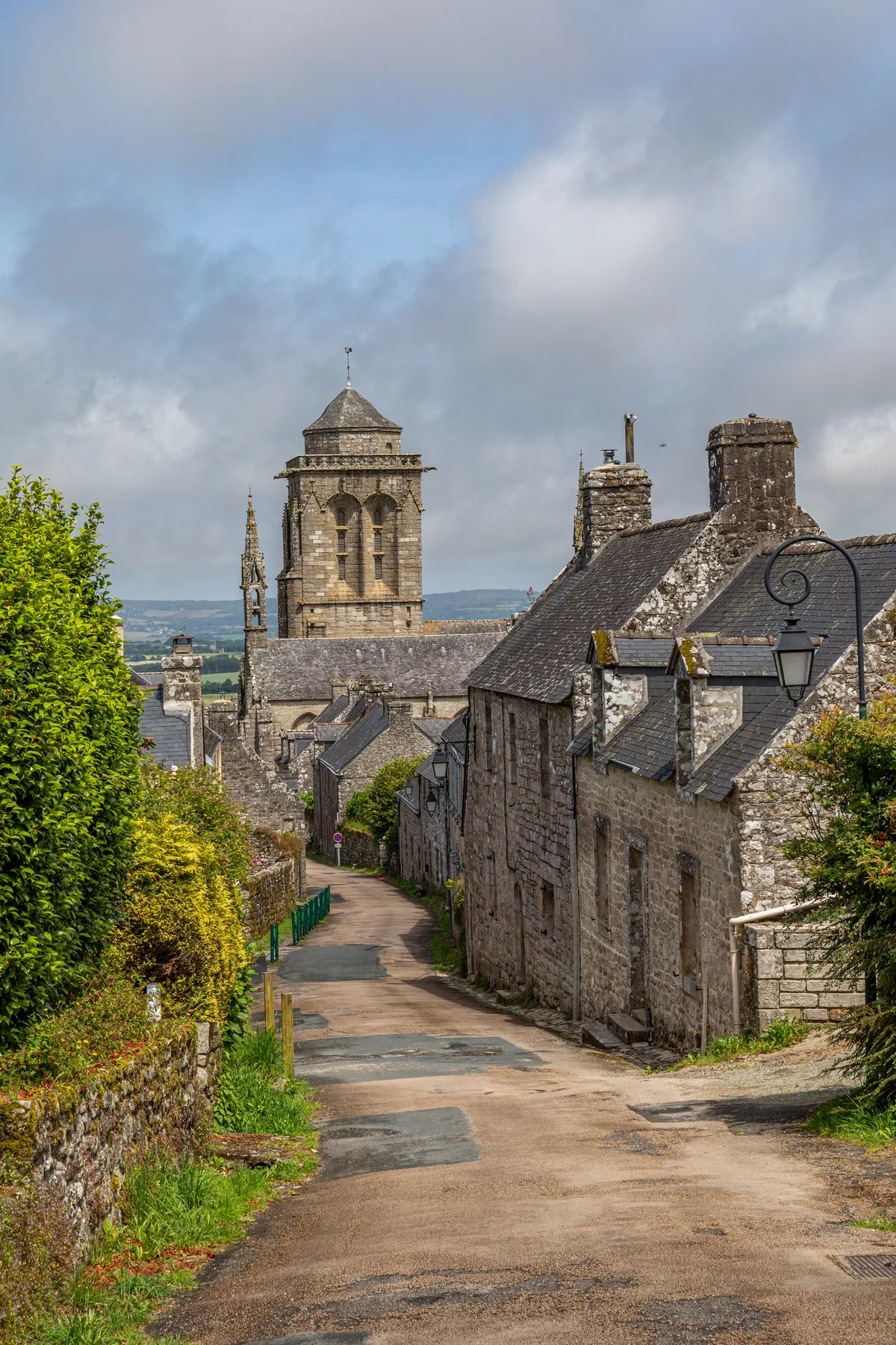 In dem malerischen französischen Dorf Locronon führt eine schmale Straße zwischen alten Steinhäusern mit Schieferdächern hinunter. In der Ferne erhebt sich ein historischer Kirchturm unter einem teilweise bewölkten Himmel. Üppiges Grün umgibt die Szene.