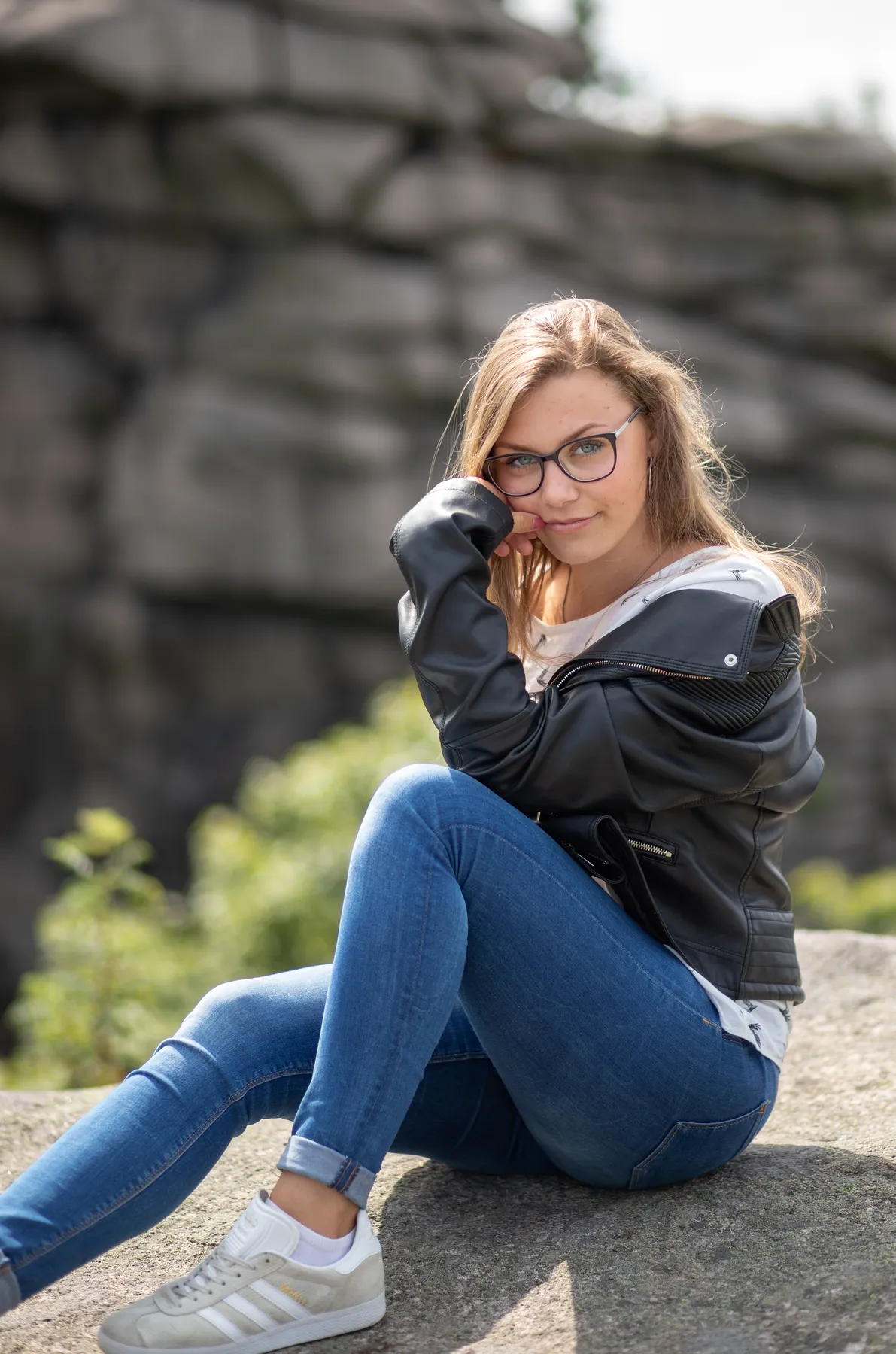 Portraitfotografie einer jungen Frau mit Brille und Lederjacke, entspannt sitzend auf einem Felsen in natürlicher Outdoor-Umgebung mit weichem Hintergrund.