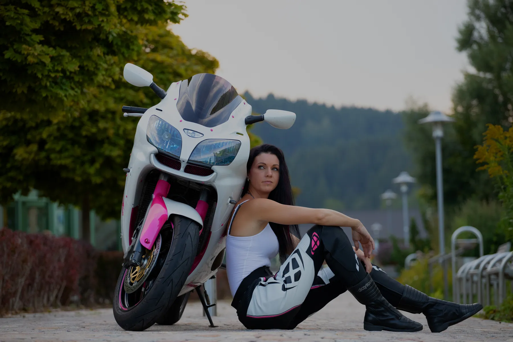 Portraitfotografie einer selbstbewussten Frau in Motorradbekleidung, sitzend neben einem Sportmotorrad in urbaner Umgebung mit natürlichem Licht.