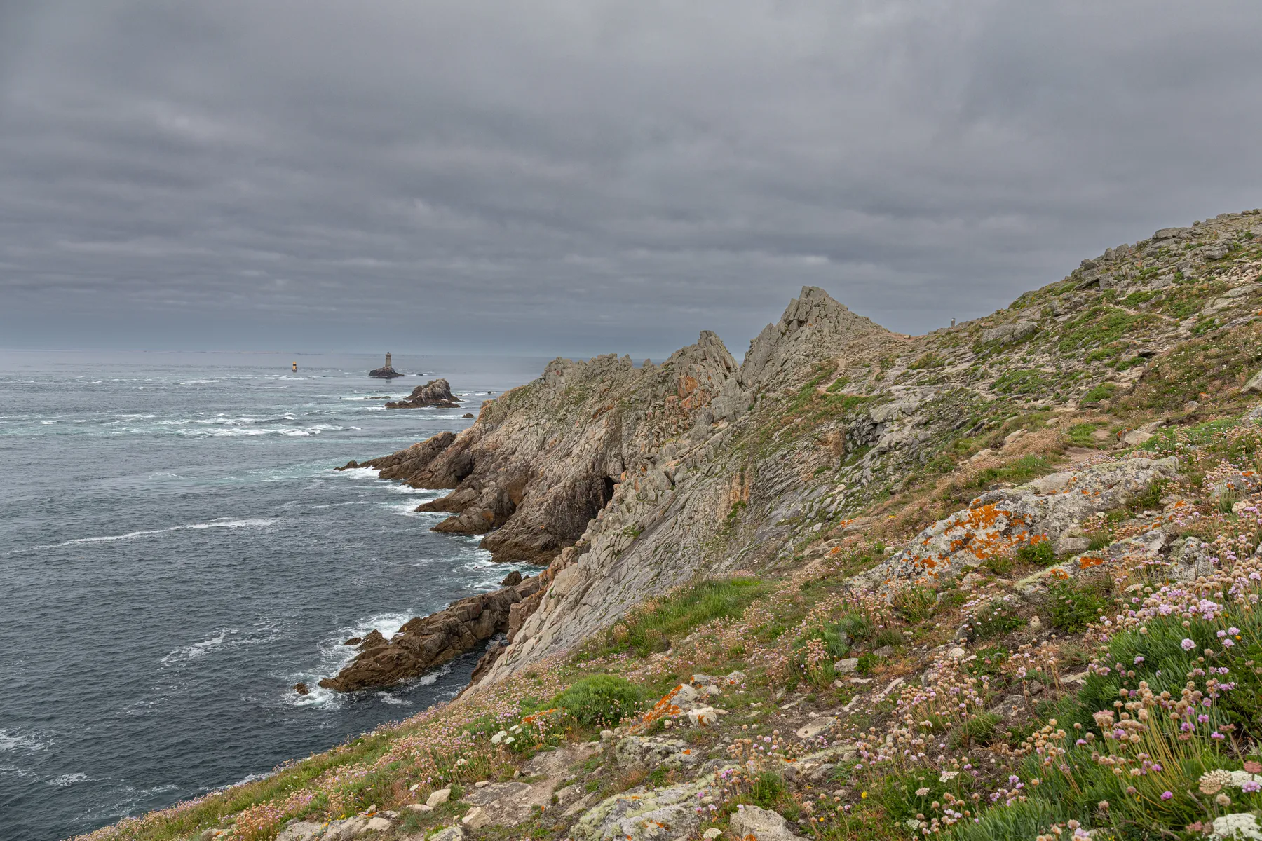 Landschaftsfotografie einer markanten Küstenlinie mit rauen Klippen im Vordergrund, Meereswellen und ein Leuchtturm auf einer entfernten kleinen Felseninsel unter einem bewölkten Himmel.