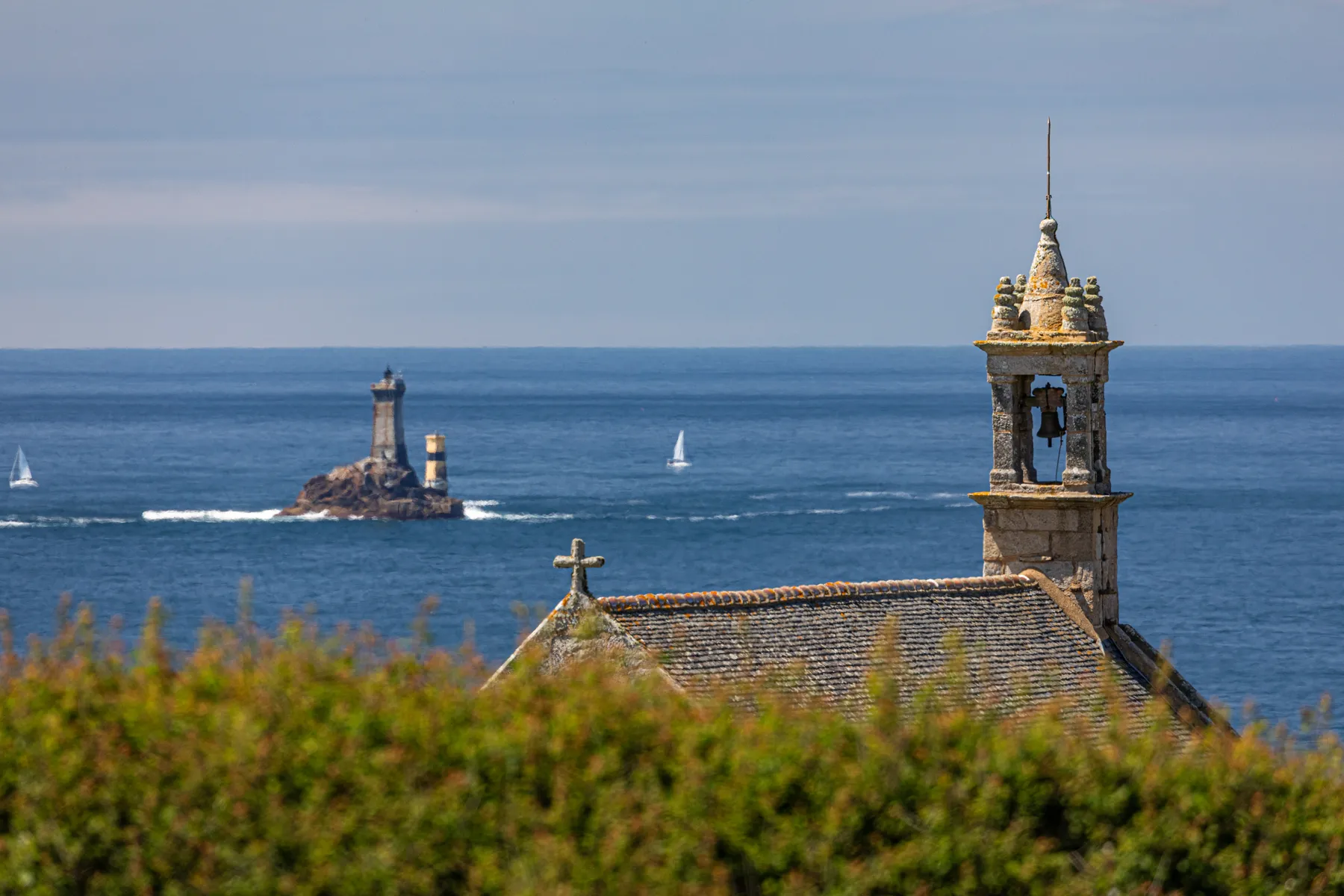 Im Vordergrund steht ein steinerner Kirchturm mit einem Kreuz auf dem Dach. In der Ferne erhebt sich ein Leuchtturm auf einer felsigen Insel im blauen Meer unter einem teilweise bewölkten Himmel, mit einigen Segelbooten auf dem Wasser.