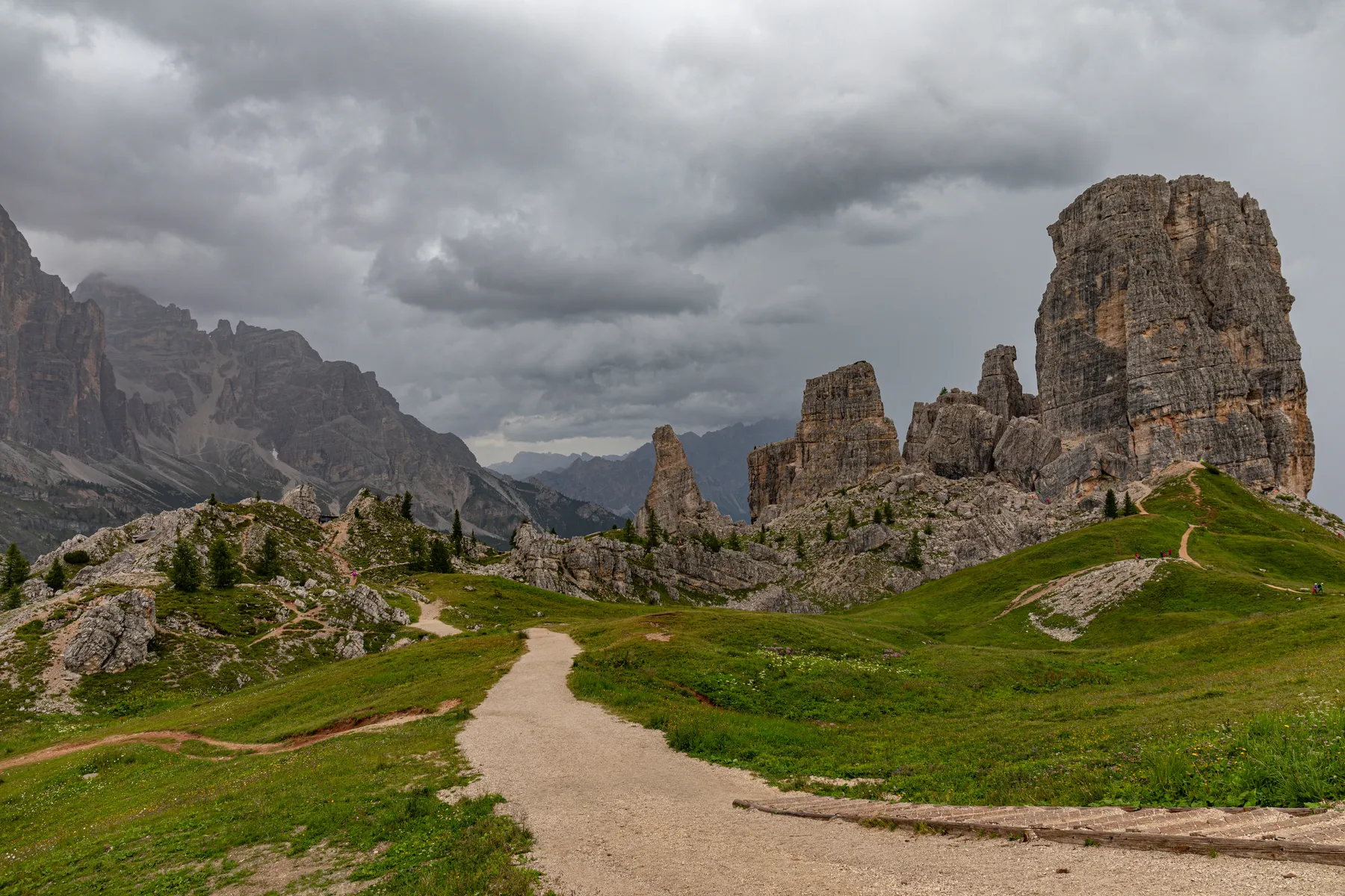 Ein gewundener Wanderweg führt durch grüne Hügel zu dramatischen, zerklüfteten Felsformationen unter einem wolkenverhangenen, grauen Himmel in einer bergigen Landschaft der Dolomiten