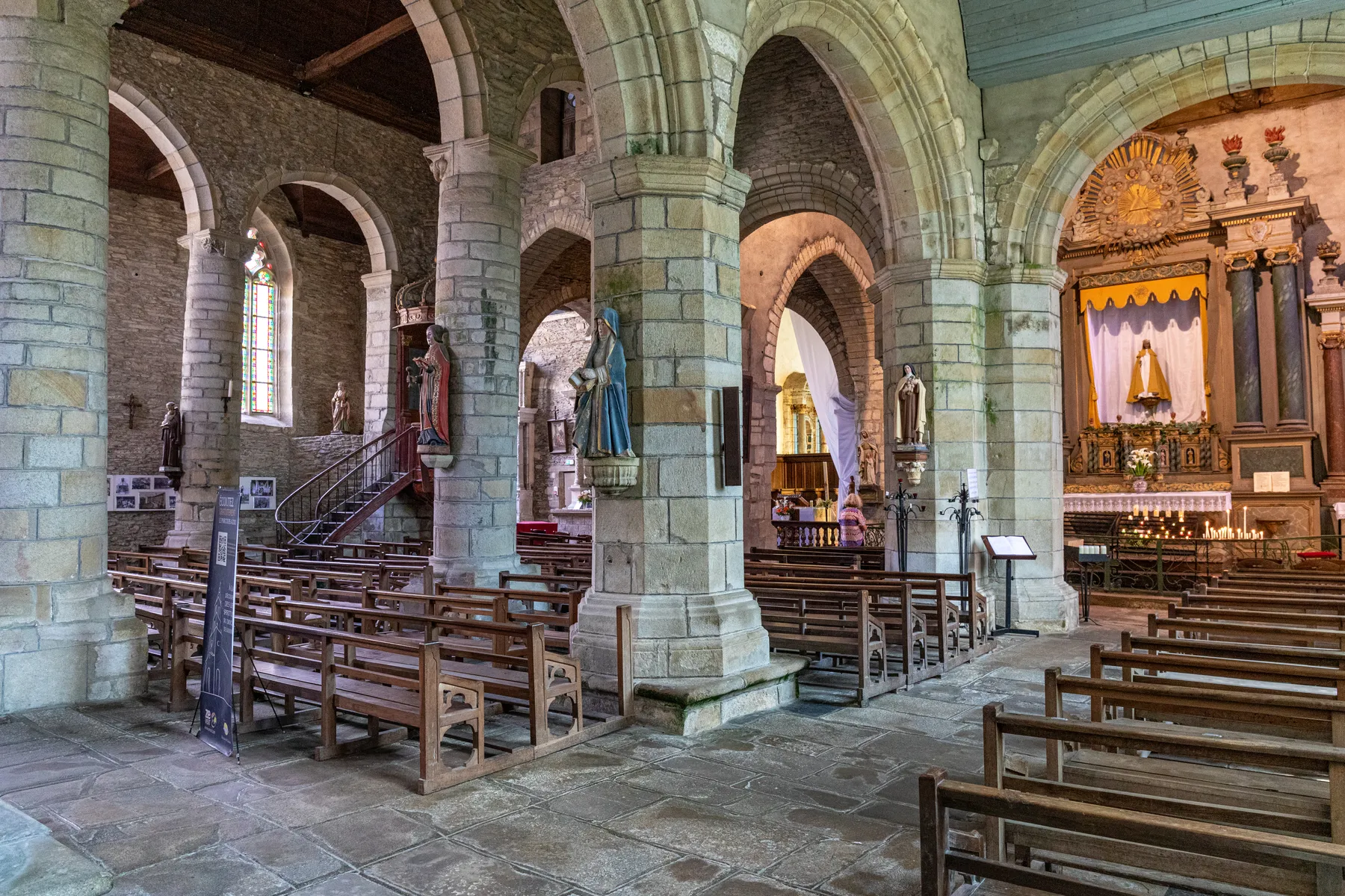 Das Innere einer historischen Steinkirche mit hölzernen Kirchenbänken, gewölbten Säulen, religiösen Statuen, Buntglasfenstern und einem mit Kerzen und religiösen Ikonen geschmückten Altar.