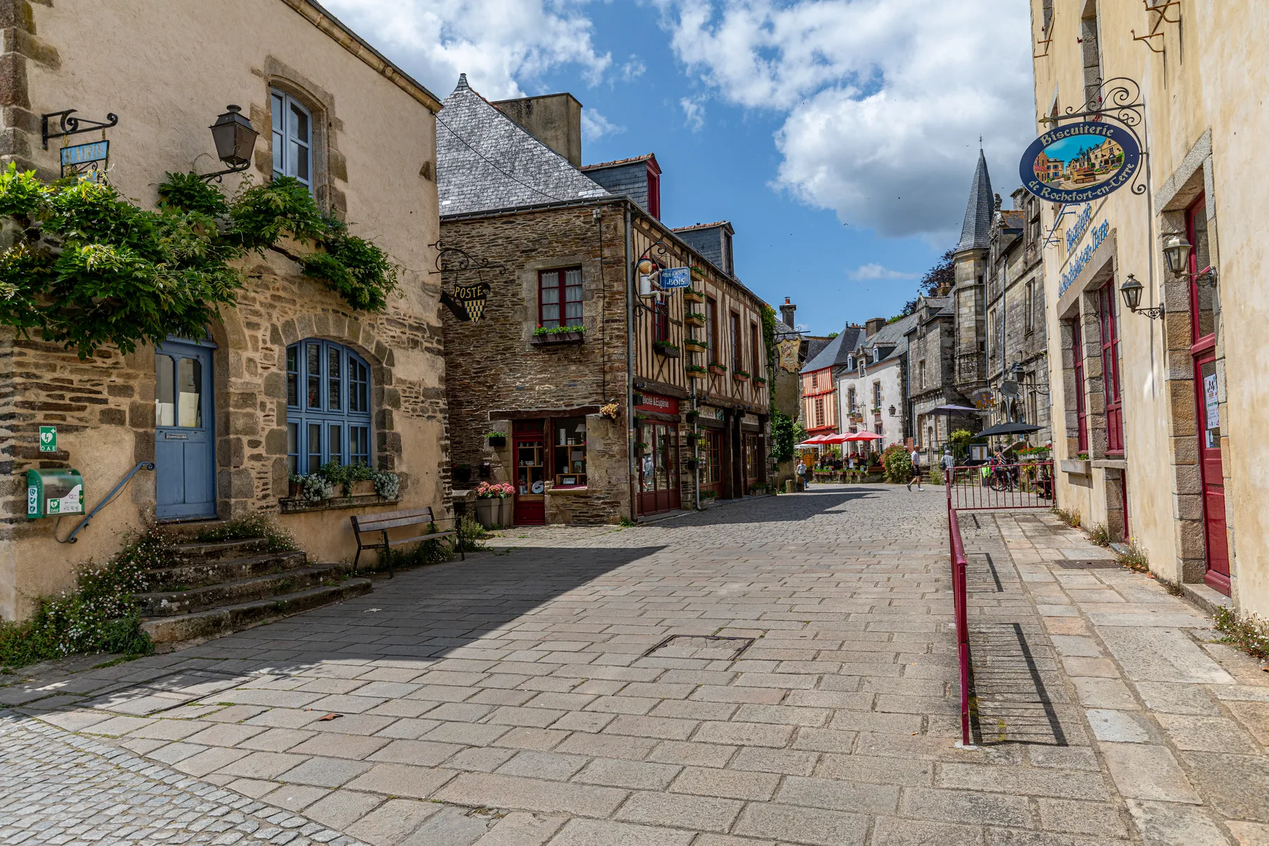 Eine ruhige Kopfsteinpflasterstraße in dem französischen Dorf Rochefort-en-Terre, gesäumt von historischen Steingebäuden, bunten Schaufenstern, Blumen und einem blauen Himmel mit vereinzelten Wolken darüber.