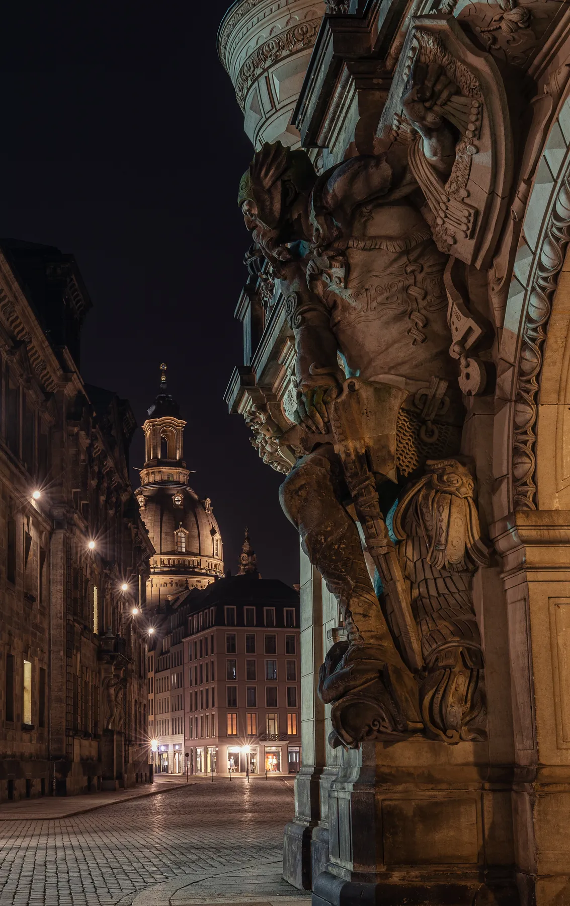 Dresden: Eine nächtliche Straßenansicht mit beleuchteten historischen Gebäuden, einer detaillierten Steinskulptur einer gepanzerten Figur im Vordergrund und der beleuchteten Kuppel der Frauenkirche im Hintergrund.