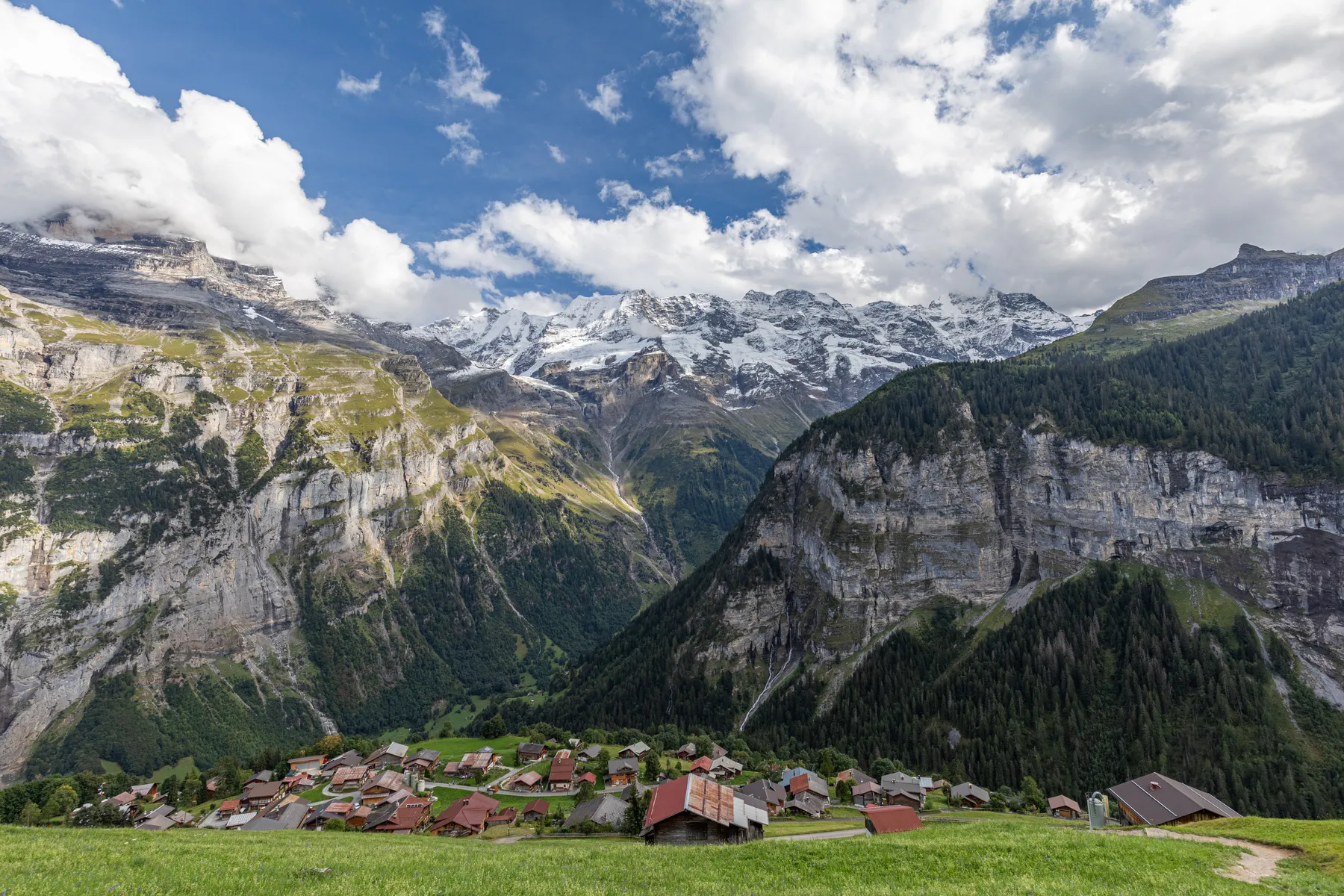 Gimmelwald ist ein kleines autofreies Dorf mit roten Dächern, eingebettet in ein grünes Tal, umgeben von steilen Felsen und hohen, schneebedeckten Bergen unter einem teilweise bewölkten Himmel.
