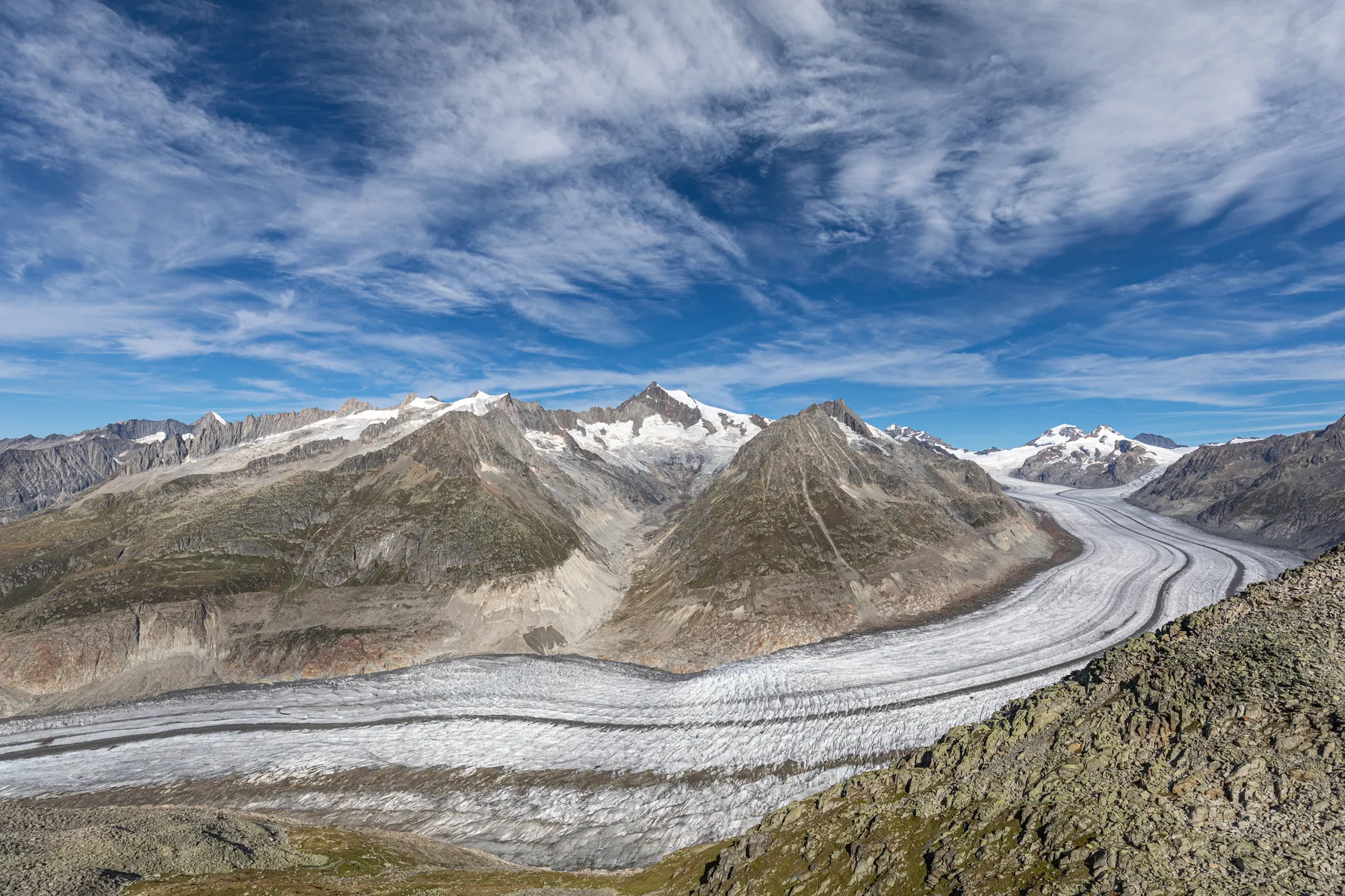 Panoramablick auf den Aletschgletscher, der sich unter blauem Himmel mit Wolkenfetzen durch ein Bergtal schlängelt, umgeben von felsigen Hängen und schneebedeckten Gipfeln.
