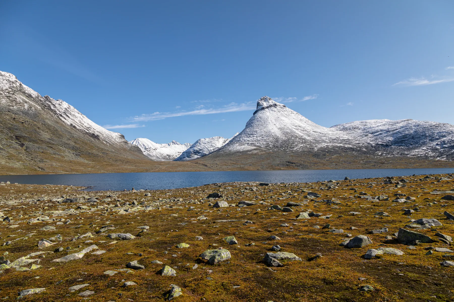 Schneebedeckte Berge umgeben einen ruhigen See unter strahlend blauem Himmel, mit einem felsigen, moosbewachsenen Vordergrund und verstreuten Steinen.
