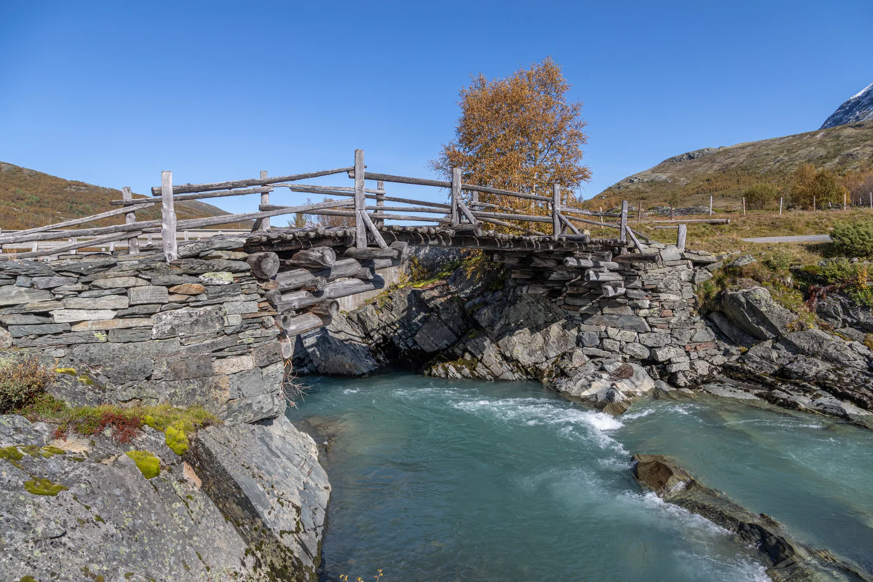 Landschaftsfotografie einer rustikalen Brücke aus Holz und Stein überquert einen klaren, blauen Bach in einer felsigen, bergigen Landschaft. Ein goldener Herbstbaum steht hinter der Brücke unter einem klaren blauen Himmel.