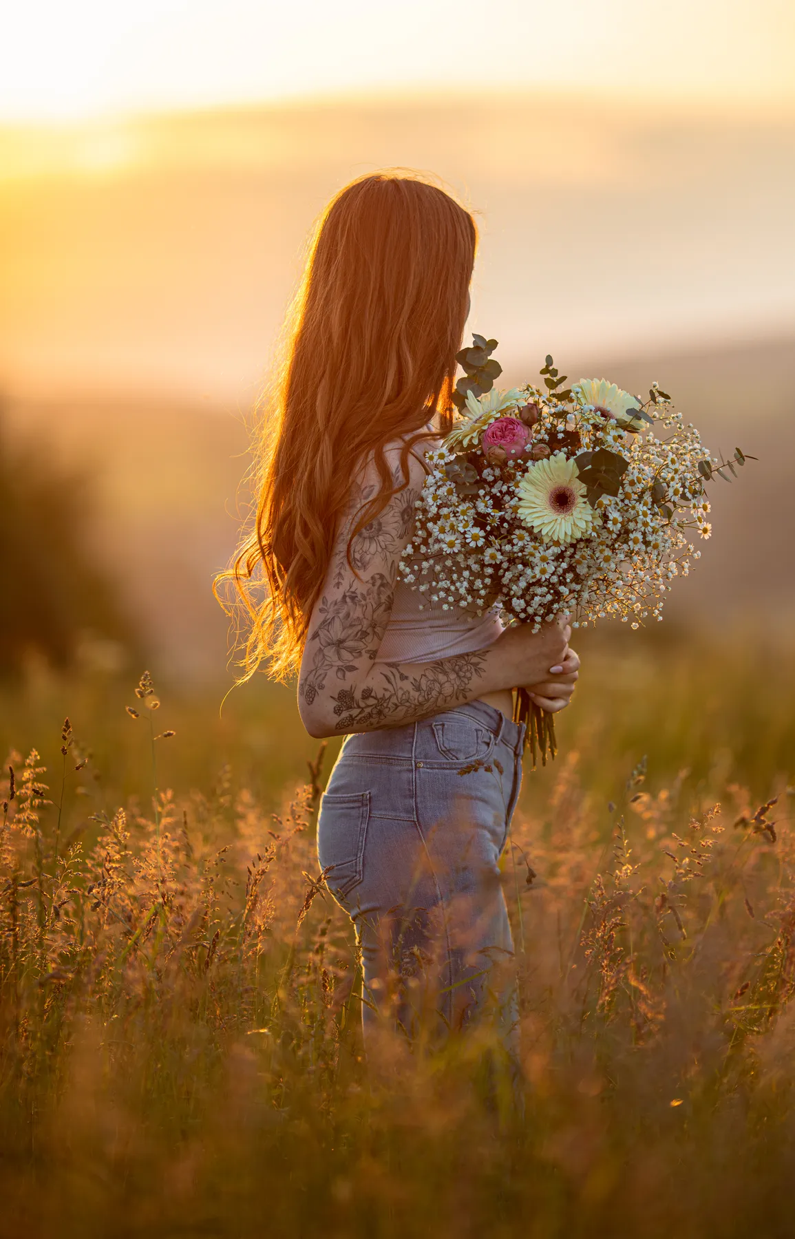 Eine Frau mit langen roten Haaren und Tätowierungen steht in einem sonnenbeschienenen Feld, hält einen Blumenstrauß in der Hand und blickt von der Kamera weg in Richtung eines goldenen Sonnenuntergangs.