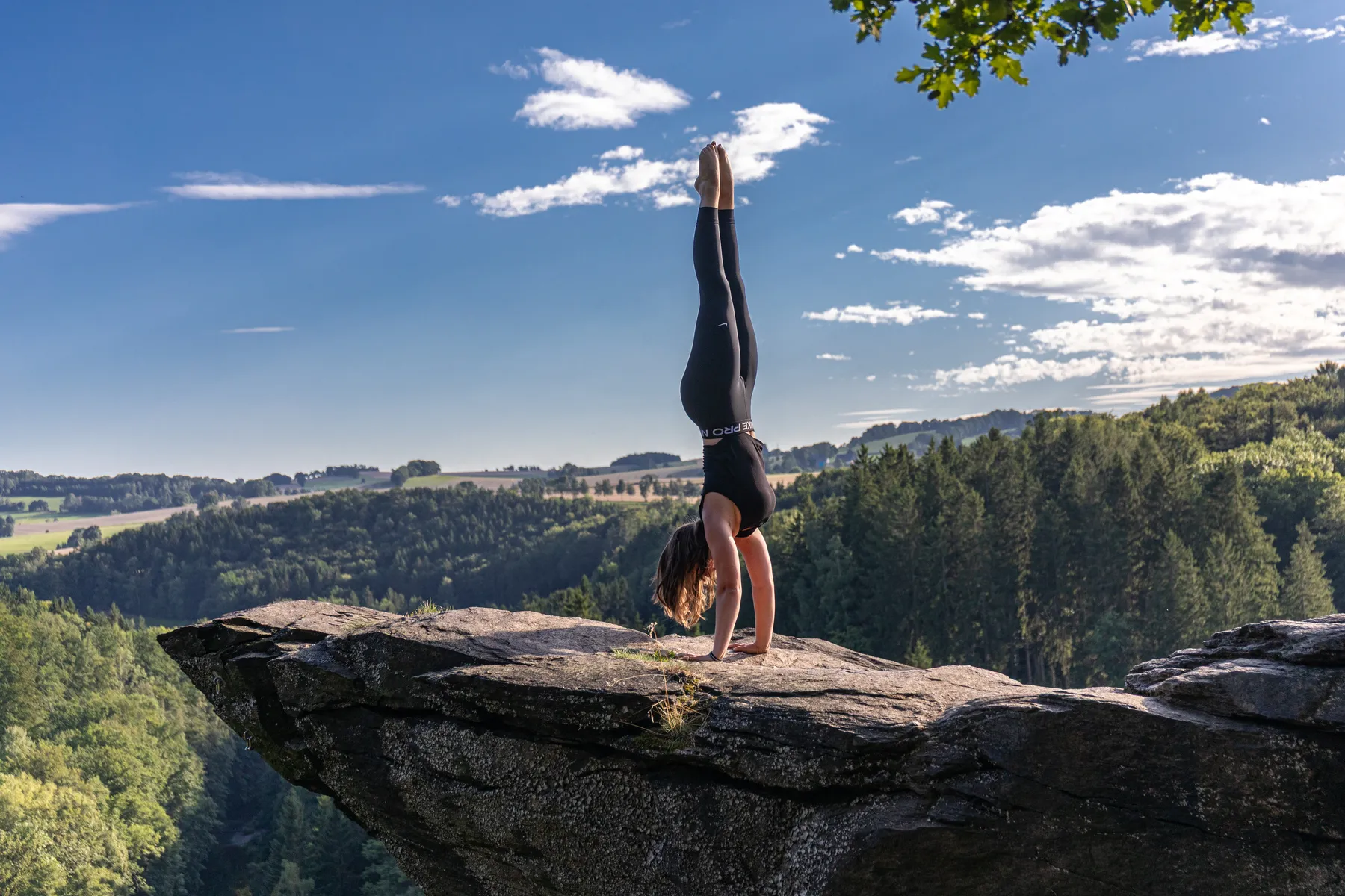 Sportfotografie einer Turnerin in schwarzer Sportkleidung die einen Handstand mit gestreckten Beinen auf einem großen Felsvorsprung macht. Im Hintergrund eine bewaldete Landschaft unter einem teilweise bewölkten Himmel.