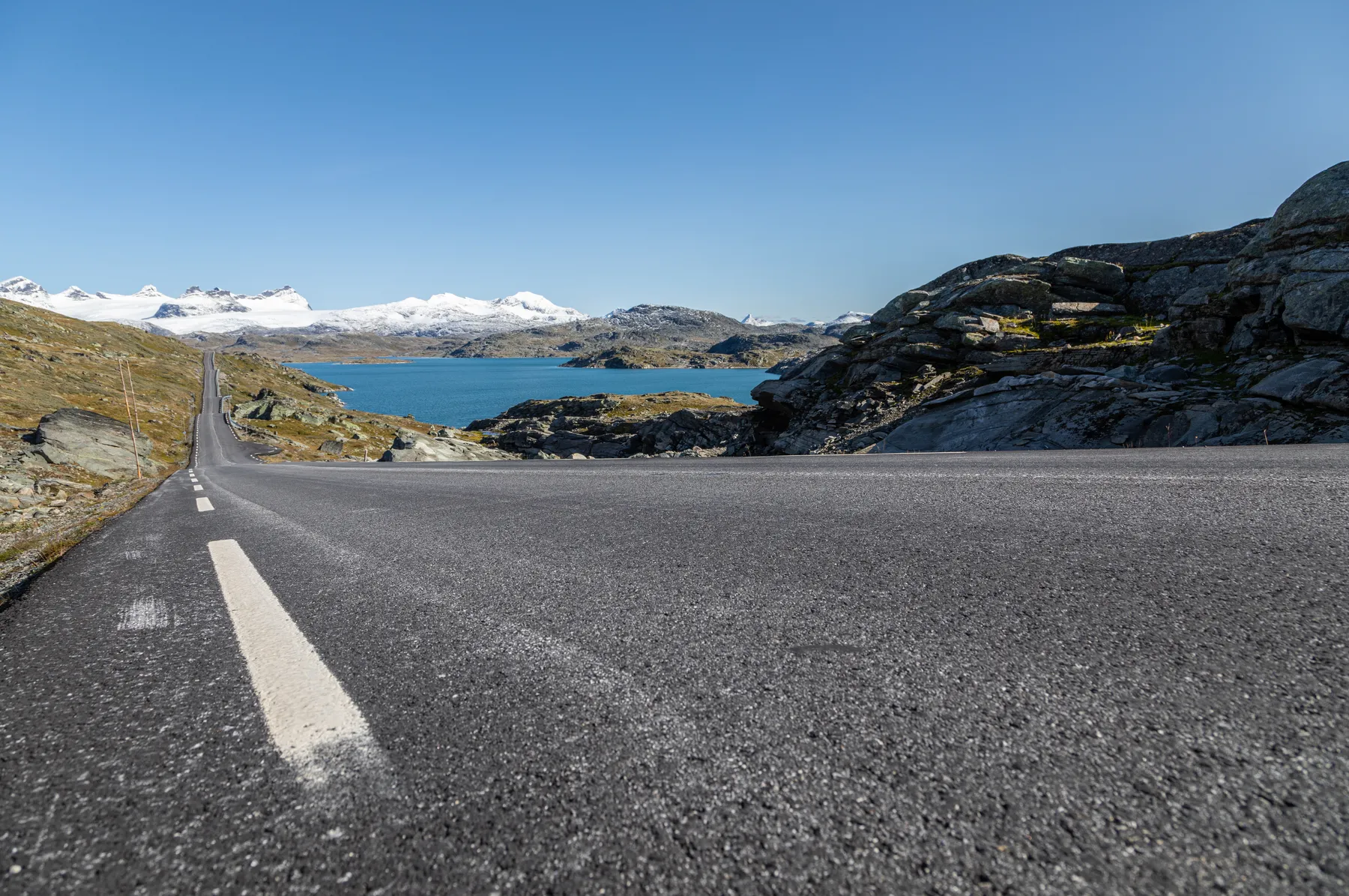 Eine asphaltierte Straße erstreckt sich in der Ferne neben felsigen Hügeln und einem blauen See, mit schneebedeckten Bergen unter einem klaren Himmel im Hintergrund.