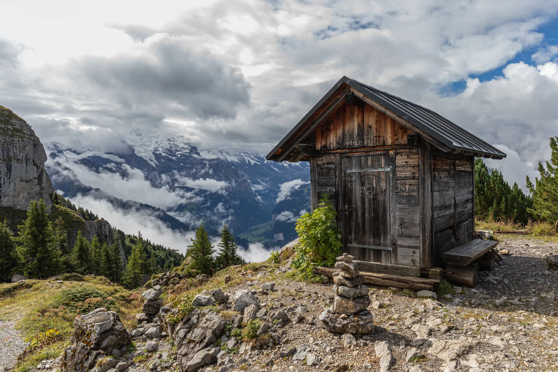 Eine kleine, rustikale Holzhütte liegt auf einem felsigen Hügel mit Kiefern in der Nähe, mit Blick auf ein Tal und die fernen schneebedeckten Berge, die teilweise von Wolken bedeckt sind. Dichter Nebel hängt an den Bergwänden im Hintergrund