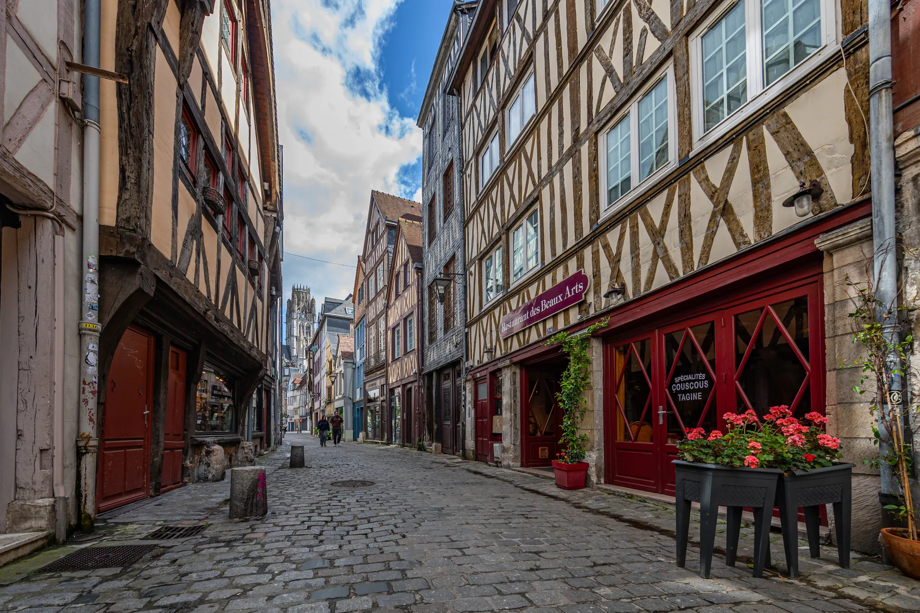 Mittelalterliche Gasse mit Fachwerkhäusern zu beiden Seiten, man sieht zqwi Menschen in der Gasse un dim Hintergrund erhebt sich ein mittelalterlicher Kirchenbau