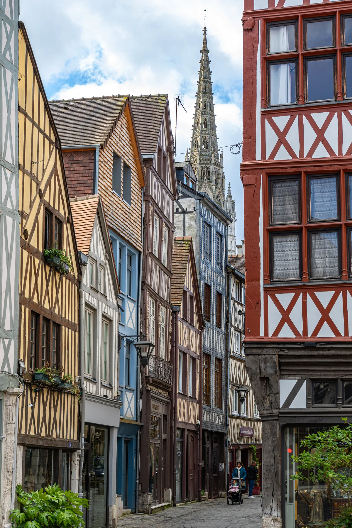 Im Bild ist eine schmale Gasse zusehen welche gesäumt ist von mittelalterlichen Fachwerkbauten, im Hintergrund erhebt sich der Turm einer großen Kirche. In der Gasse ist eine Frau mit Kinderwagen unterwegs