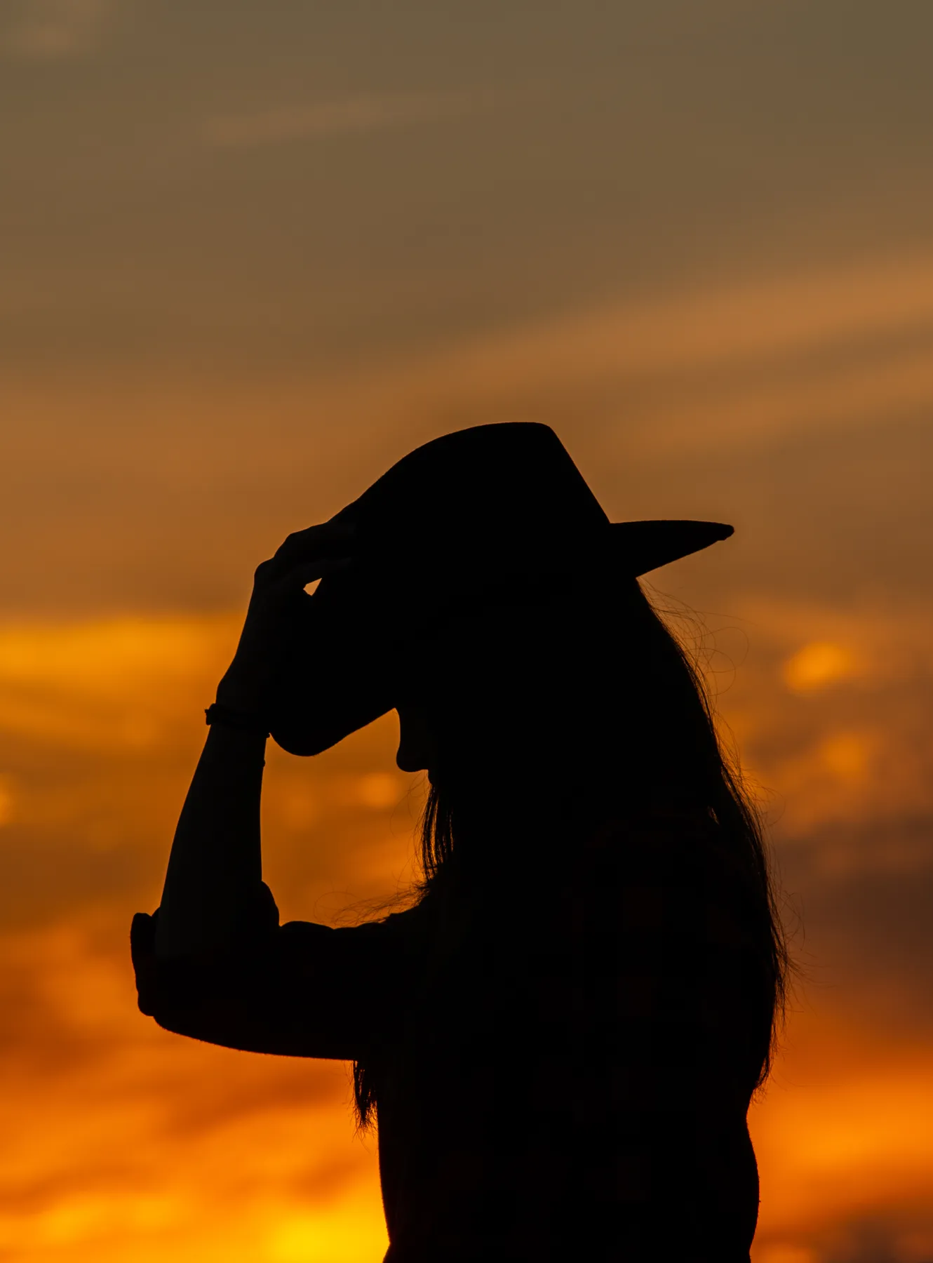 Cowgirls steht als Silhouette vor einem Sonnenuntergang und greift mit einer Hand an ihren Cowboyhut