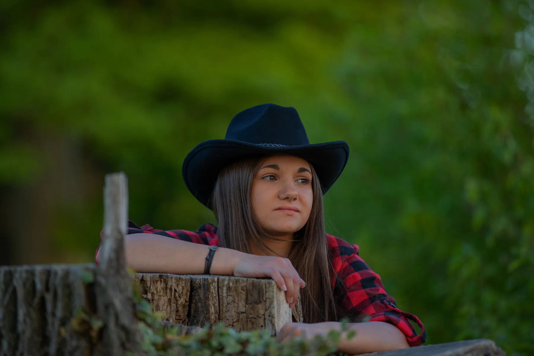 Junges Mädchen in kariertem Hemd und schwarzem Cowboyhut lehnt auf einem Baumstamm