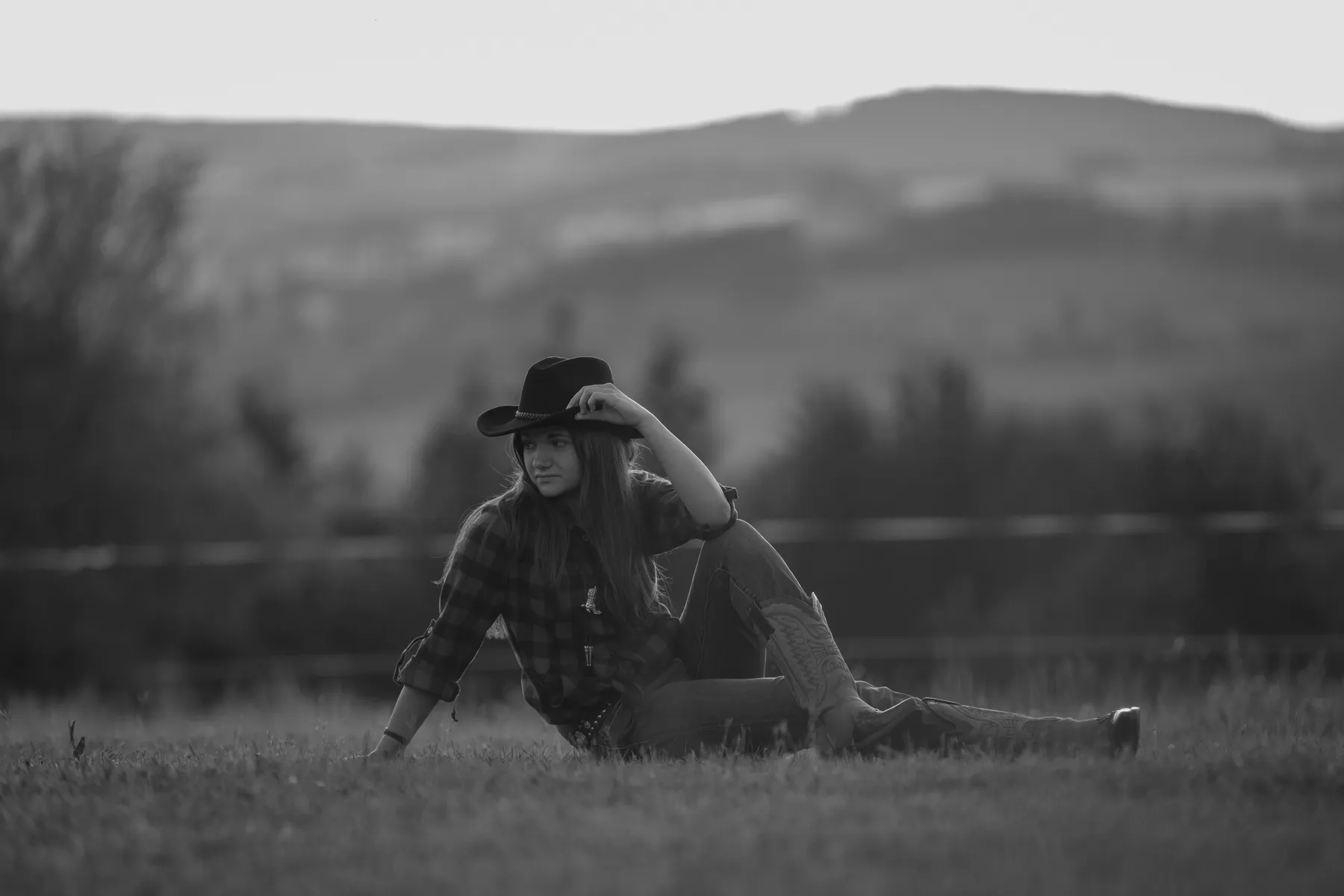 Schwarz weiß Foto eines jungen Cowgirls was auf einer Wiese sitzt, ein Bein ist angewinkelt auf dem sie ihre Hand stützt und sie schaut seitlich in die Ferne