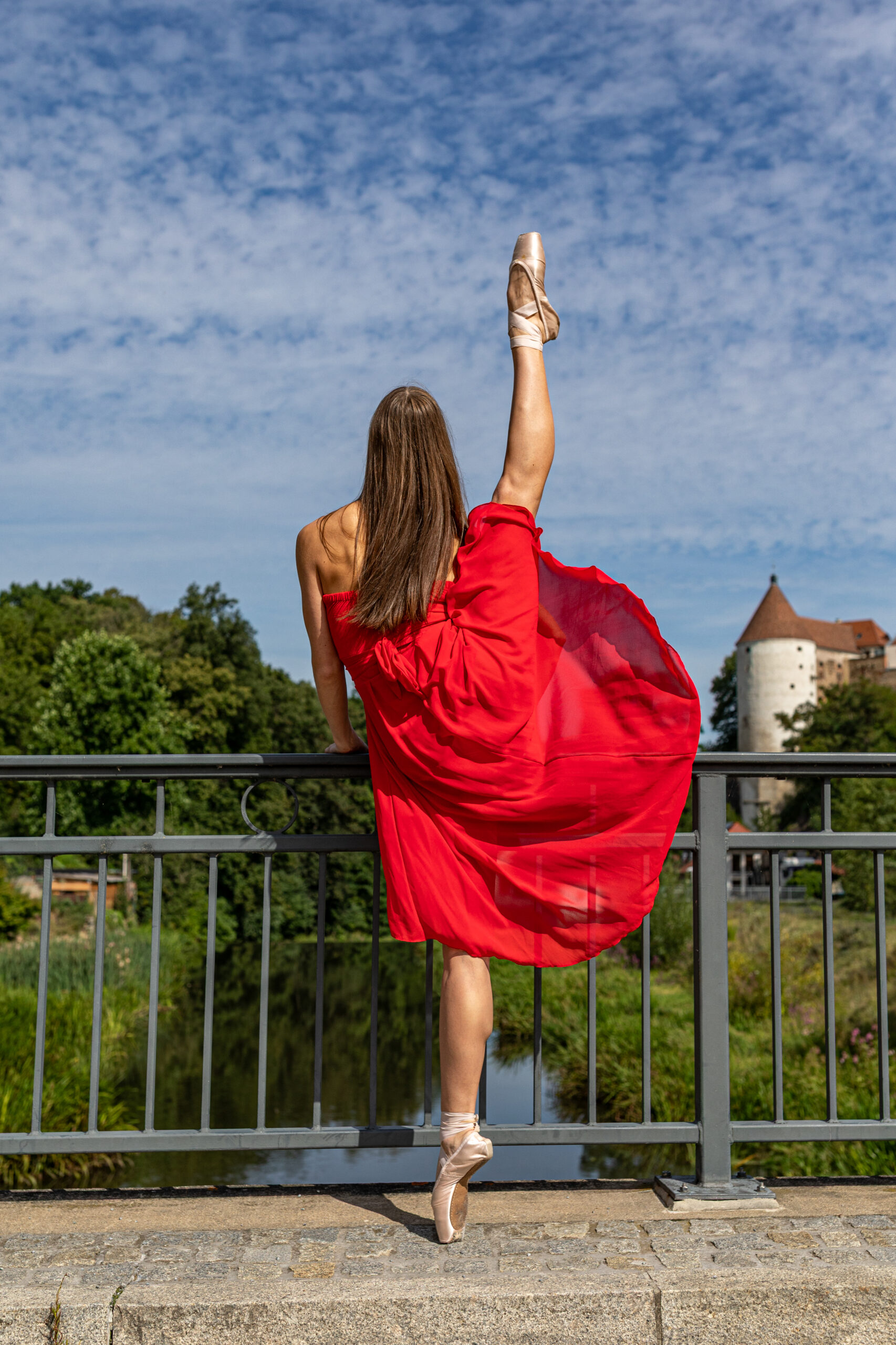 Eine Ballerina steht an einem Brückengeländer und streckt ein Bein 180 grad in die Luft, sie trägt ein rotes Kleid. Im Hintergrund ist ein Fluß und eine Burg zu sehen