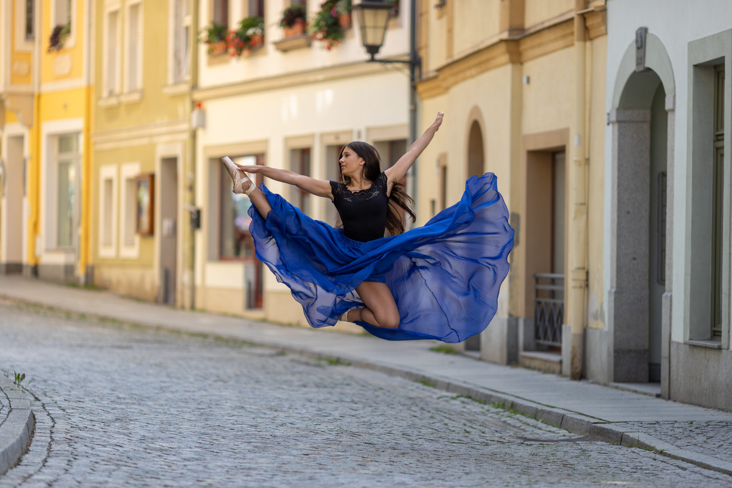 Eine Ballerina springt in einer Straße mit Kopfsteinpflaster. Sie trägt einen blauen Rock der in der Luft verweht.
