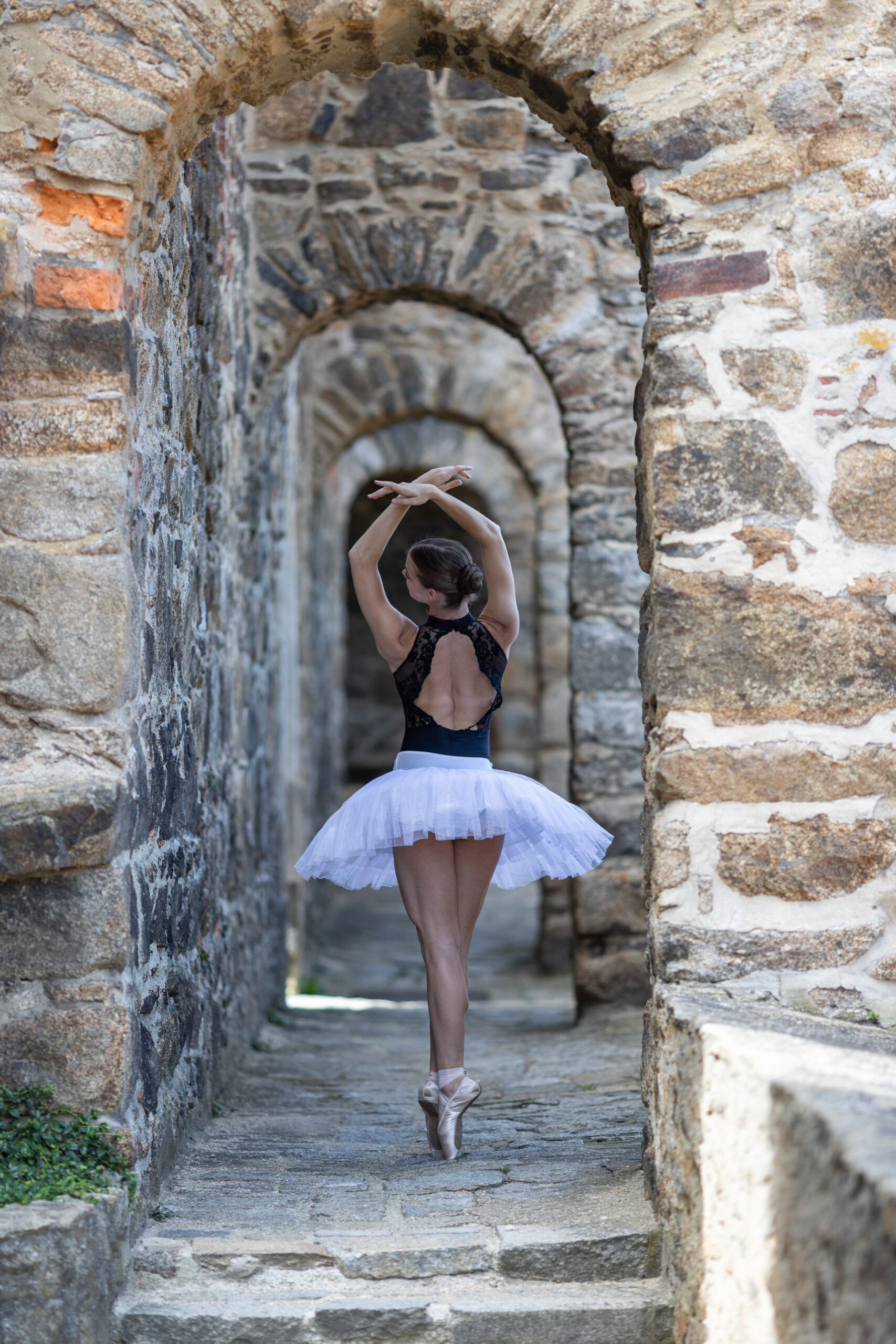 Eine Ballerina steht in einem schwarzen Body und weißen Tut auf Spitze und hält die Arme nach oben. Sie befindet sich in einem Bogengang einer mittelalterlichen Ruine