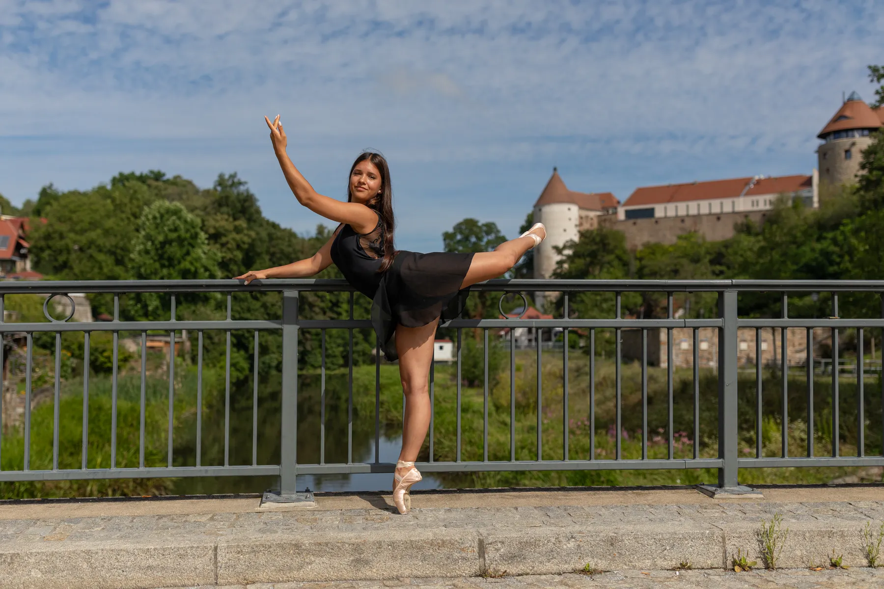Eine Ballerina steht an einem Brückengeländer, mit dem einen Bein steht sie auf der Spitze, das andere ist nach hinten gestreckt. Im Hintergrund ist ein Fluß zu sehen und Türme einer alten Stadt