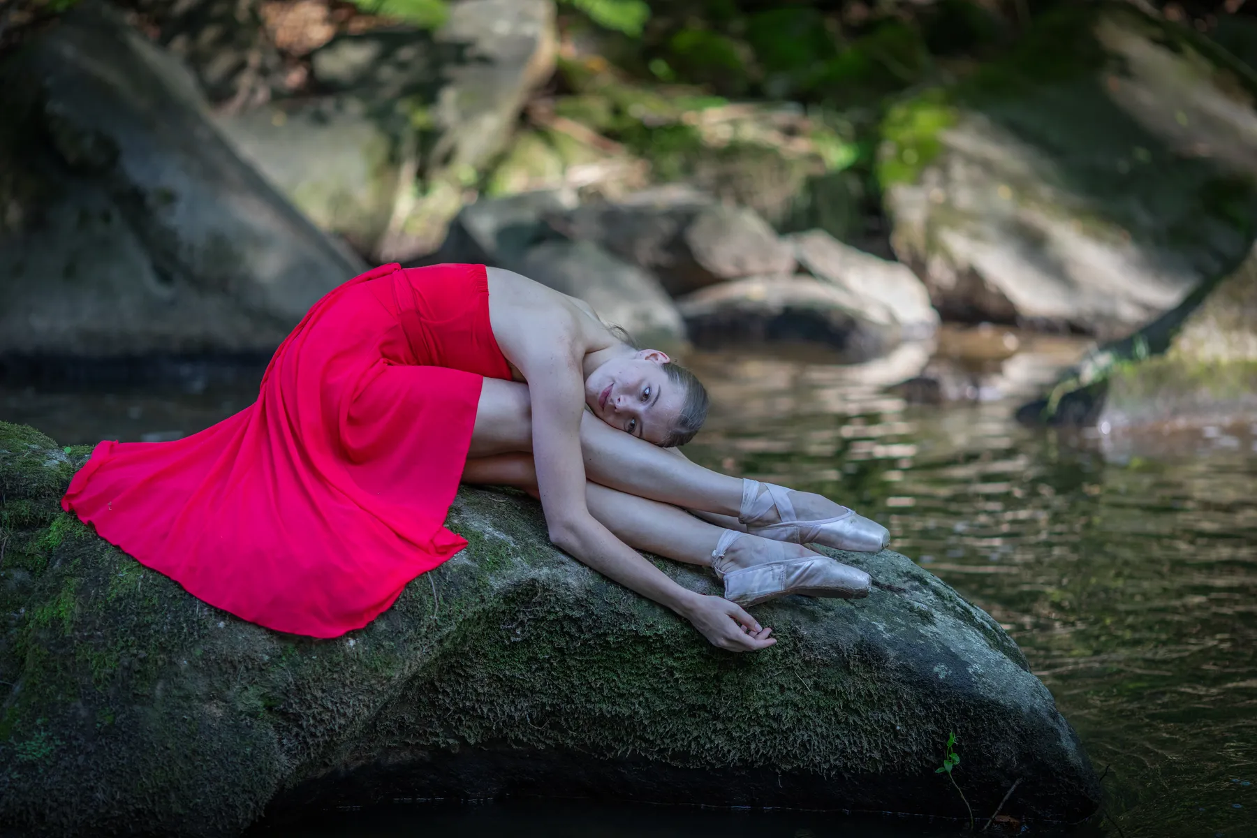 Eine Ballerina in rotem Kleid liegt in einer ruhigen Pose auf einem bemoosten Felsen am Wasser.