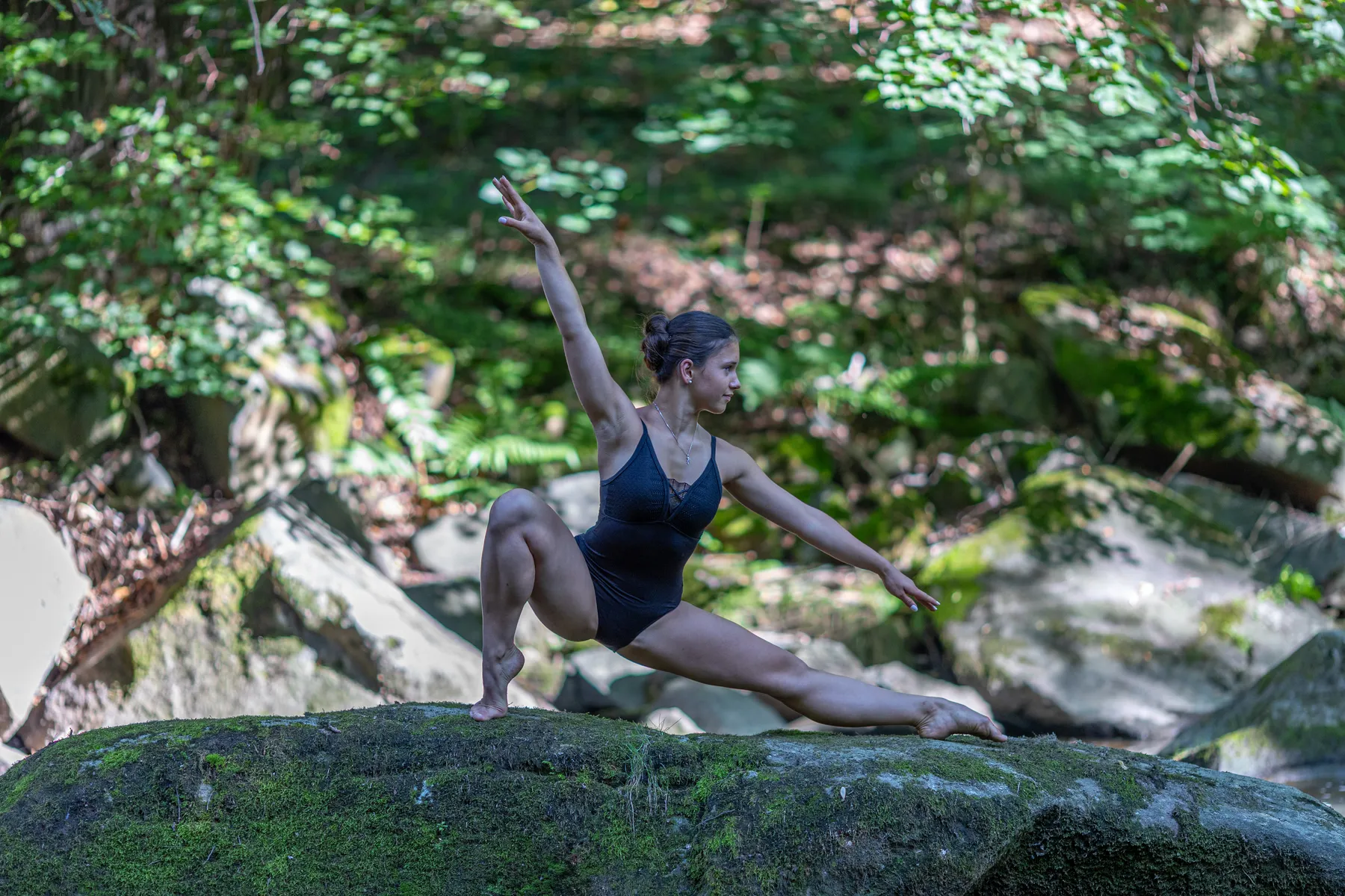 Eine Balletttänzerin zeigt eine weit ausladende Pose auf einem großen bemoosten Felsen inmitten der Natur.